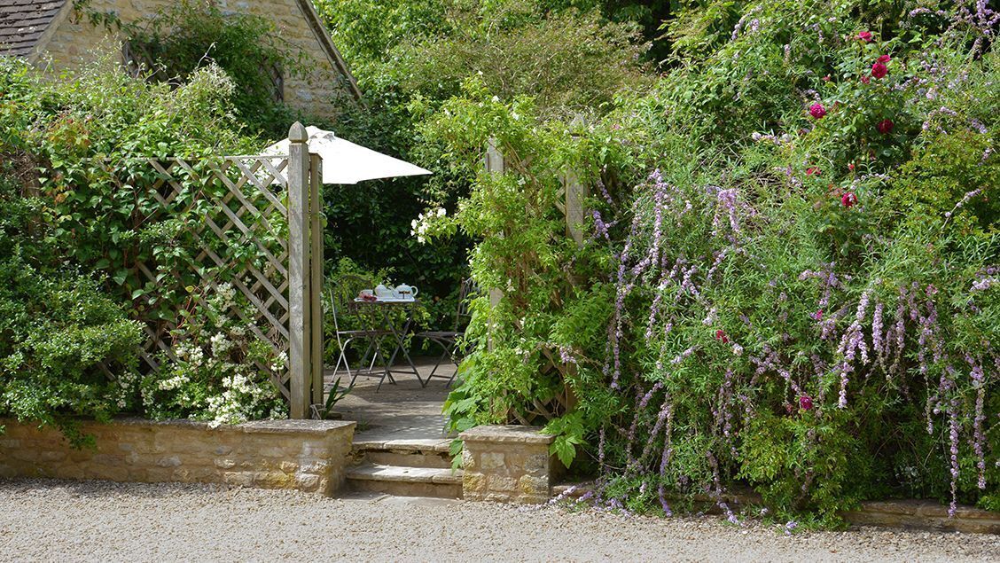 Ein Garten mit einem hölzernen Tor, einer Steinmauer und einem Kiesweg, der zu einem Tisch und Stühlen führt.