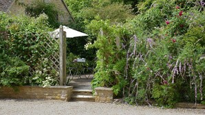 Un jardin avec une porte en bois, un mur de pierre et un chemin de gravier menant à une table et à des chaises.