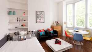 Una habitación moderna con una cama, una mesa de centro blanca, un sofá negro con cojines coloridos, una mesita de noche de madera y una mesa redonda con sillas. Se observan estanterías con objetos decorativos y una ventana con persianas.