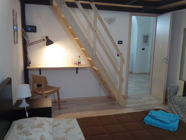 A bedroom with a bed, a desk with a chair, a wooden staircase, and a door leading to another room.
