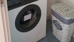 Un lavadero pequeño con lavadora de carga frontal, un cubo de basura y un calentador de agua en la pared.
