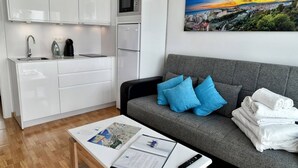Habitación de hotel moderna con kitchenette, un sofá gris con cojines azules, una cafetera y un cuadro con una vista urbana en la pared.