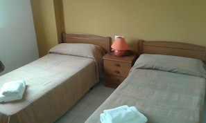 A hotel room with two single beds, a nightstand with a lamp, and a folded towel on each bed.