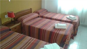 Habitación de hotel con tres camas, cada una con ropa de cama a rayas. Hay una mesita de noche con una lámpara y una pequeña mesa con una toalla doblada.