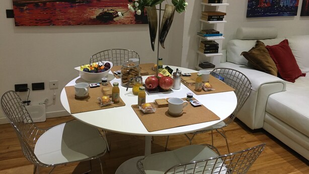 A dining area with a round table, chairs, and a variety of food items. There is a bookshelf with books and a painting of a cityscape.