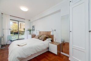 Una habitación de hotel moderna con una cama grande, mesitas de noche, un espejo y un balcón con vistas a edificios y zonas verdes.
