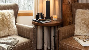 Una habitación acogedora con dos sofás a cuadros, una mesa redonda de madera con una lámpara, libros y una vela. Se ve una ventana con cortinas y vistas a árboles desnudos.