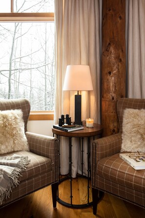 Une pièce chaleureuse avec deux canapés à carreaux, une table ronde en bois sur laquelle se trouvent une lampe, des livres et une bougie. Une fenêtre avec des rideaux offre une vue sur des arbres sans feuilles.