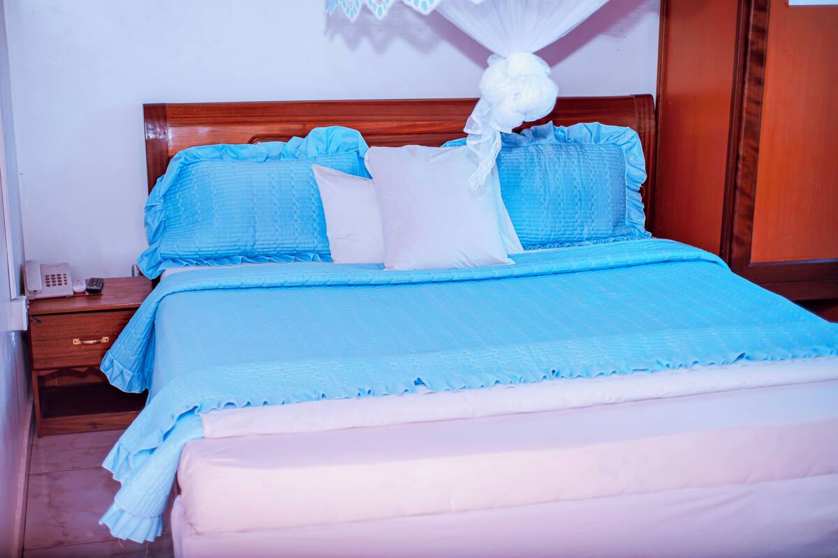 A neatly made bed with blue and white bedding, a wooden headboard, and a bedside table with a phone and a bottle.