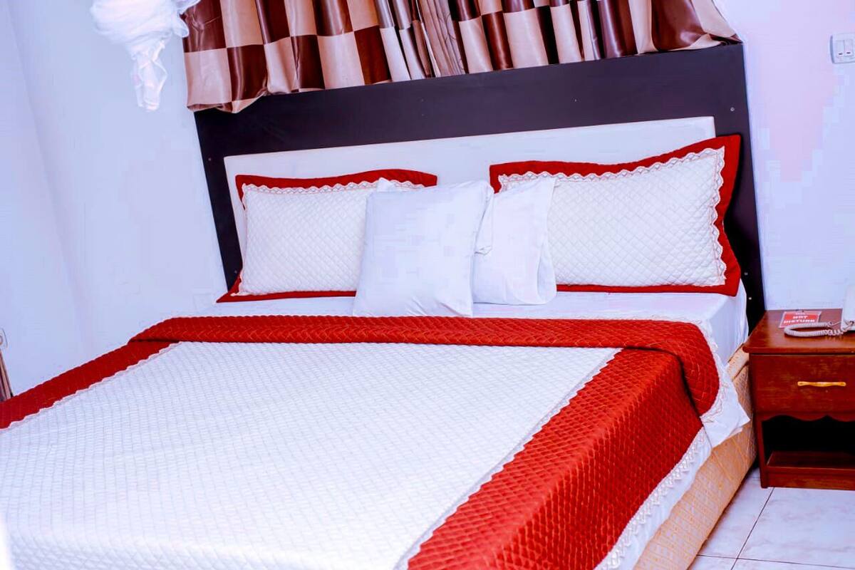 A neatly made bed with a red and white bedspread and pillows, a wooden nightstand with a book, and a curtain with a checkered pattern.