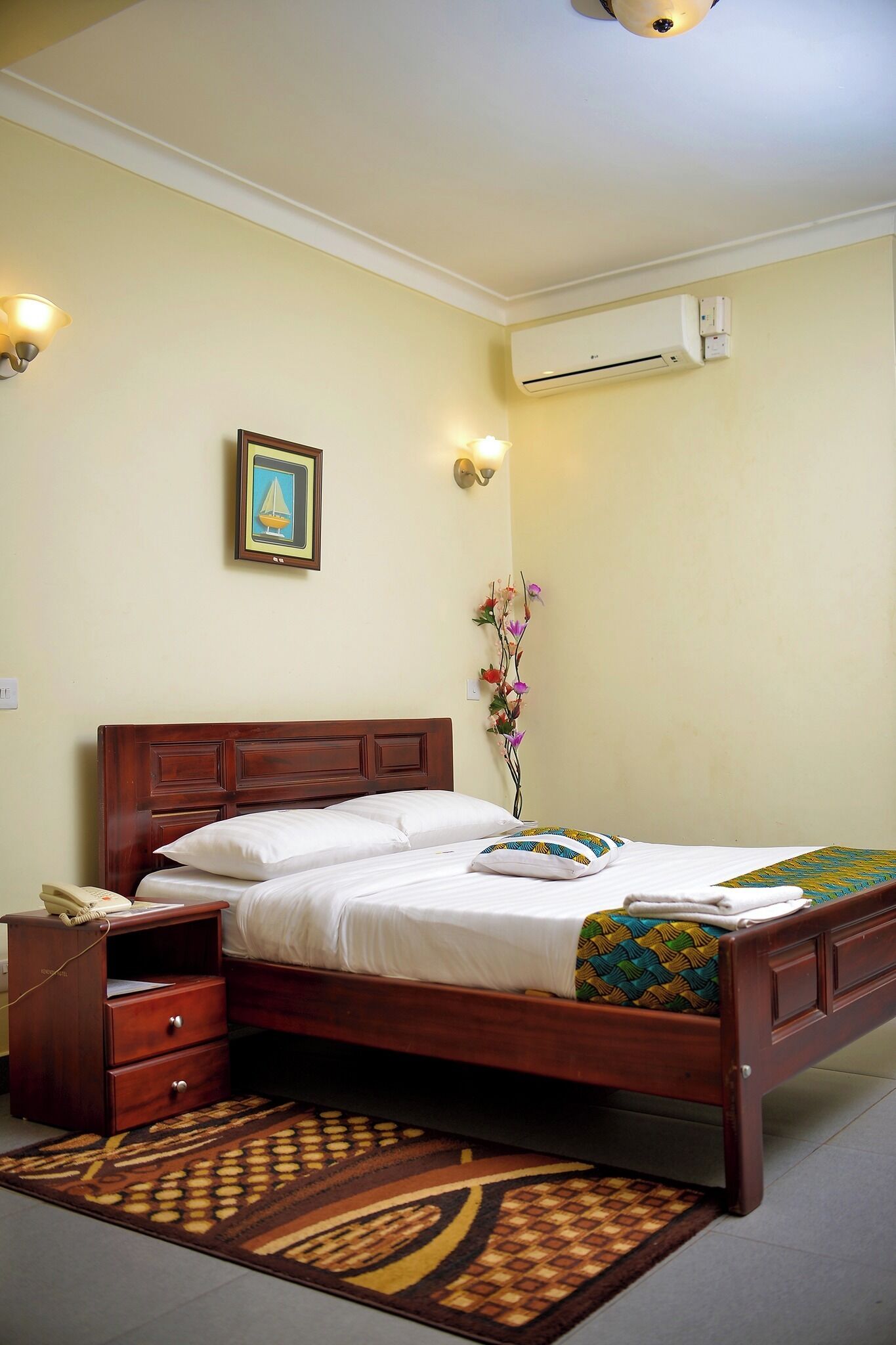 A neatly made bed with a wooden headboard, a bedside table with a lamp, and a floral arrangement on the wall.