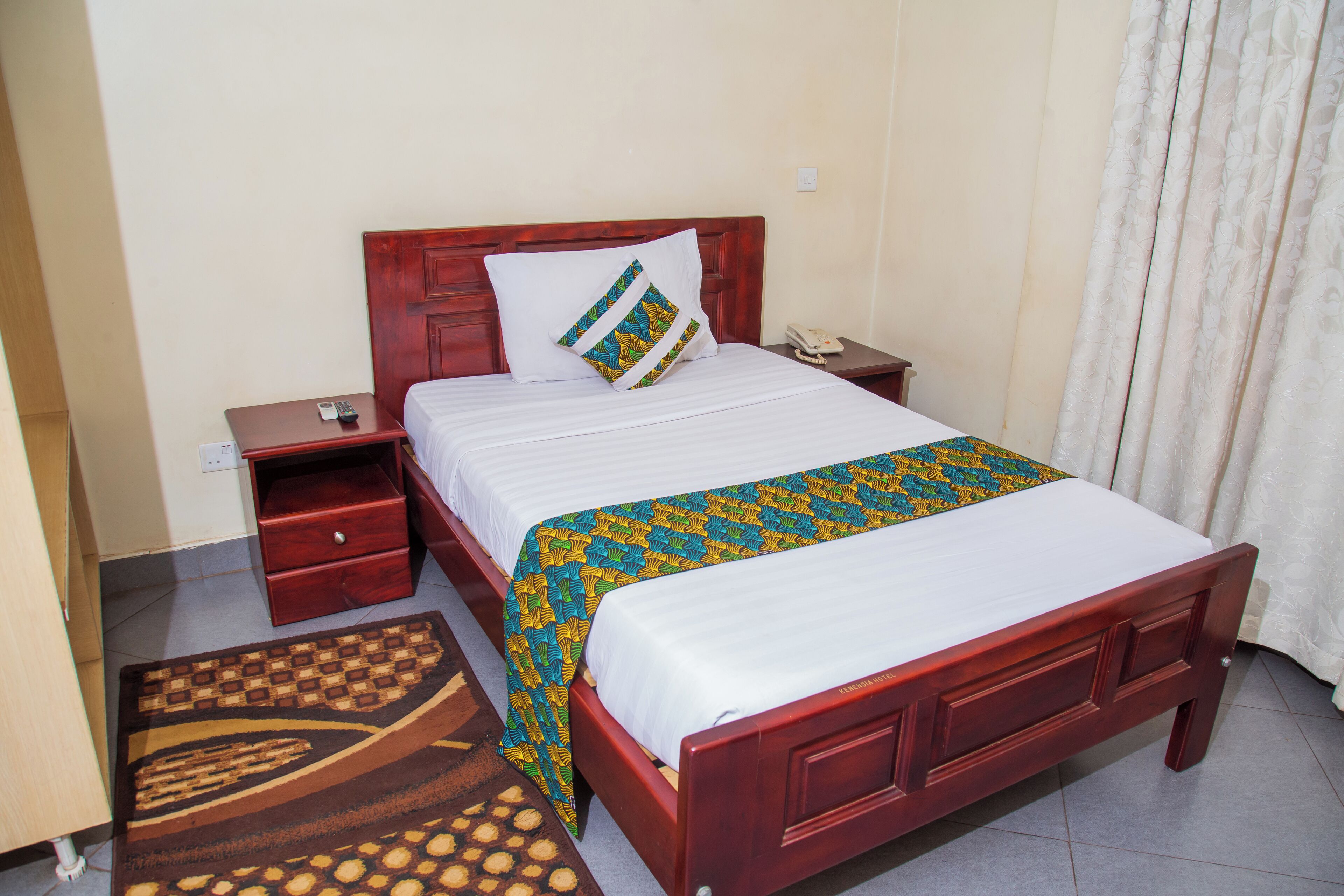 A hotel room with a wooden bed, a nightstand with a phone, a bedside table, and a patterned rug.
