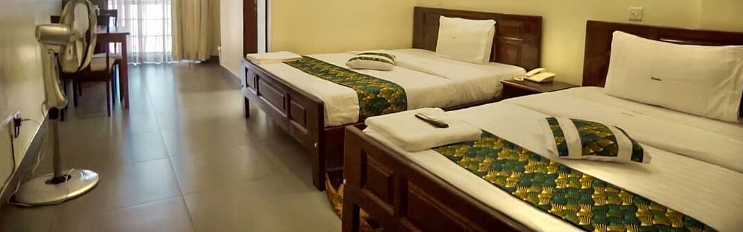 Two single beds with wooden headboards, a bedside table, a fan, and a window with curtains.