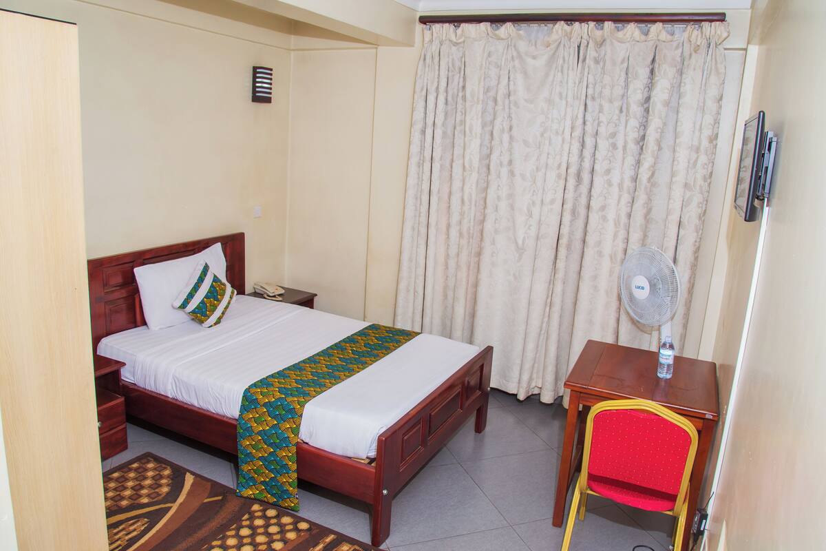 A hotel room with a bed, a desk, a chair, a fan, and a curtain.