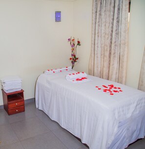 A clean, well-lit room with a bed, a small wooden side table with towels, and a decorative plant.