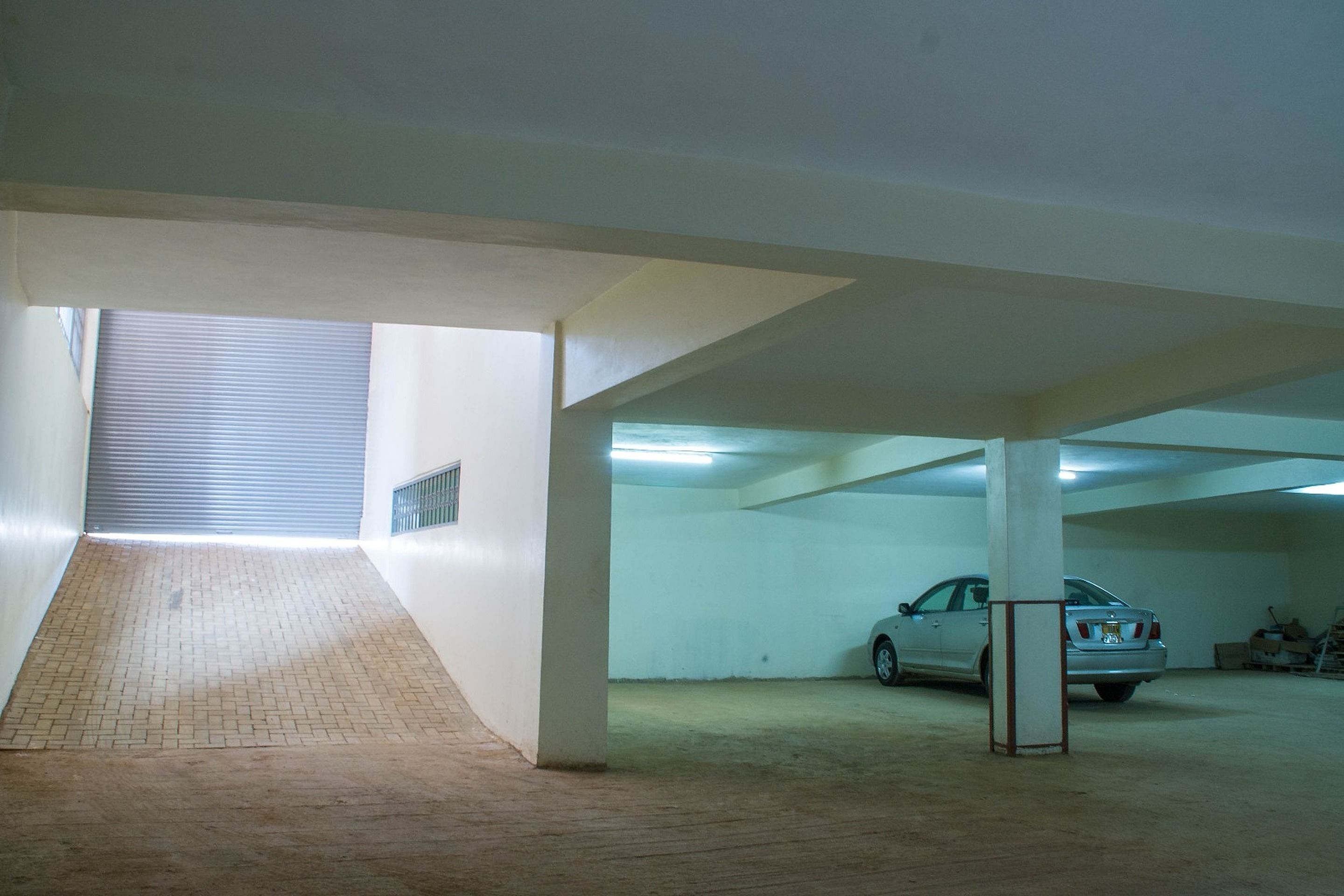 An indoor parking area with a car parked near a pillar, a ramp leading to a higher level, and a closed shutter door in the background.