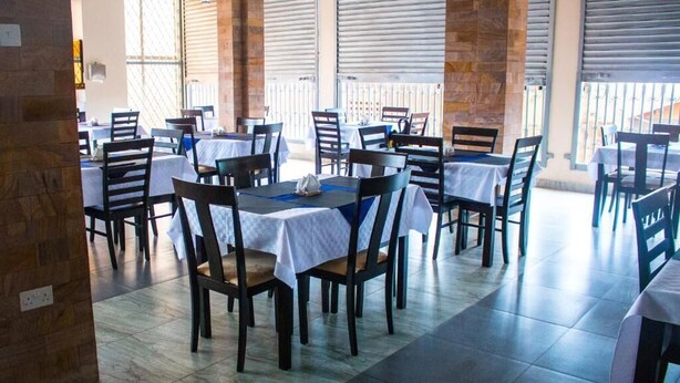 A restaurant with multiple tables and chairs, covered by a roof with metal shutters, and a stone wall on one side. - Kenendia Hotel (Kampala)