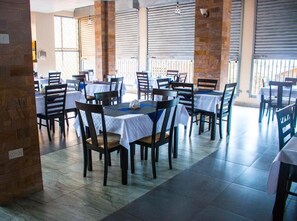 A restaurant with multiple tables and chairs, covered by a roof with metal shutters, and a stone wall on one side. - Kenendia Hotel (Kampala)