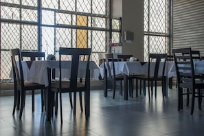 A dining area with tables and chairs, large windows with a metal grid, and a tiled floor.
