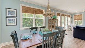 A dining area with a wooden table, chairs, and a chandelier. There are windows with blinds, a bookshelf, and a sofa in the background.