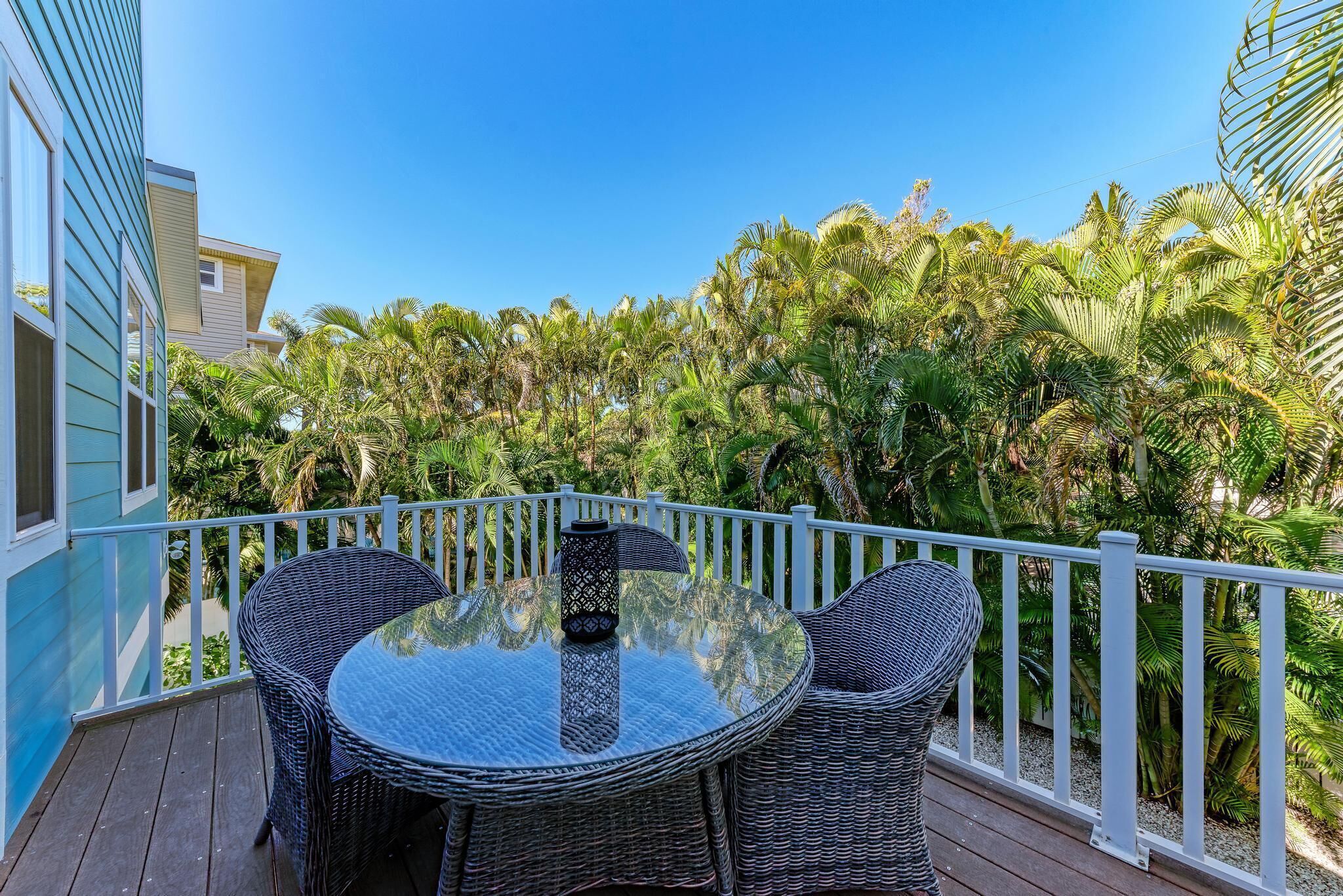Un balcon agrémenté de meubles en osier et d’une table en verre, entouré d’une balustrade blanche et de plantes tropicales luxuriantes.