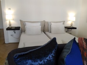 A double bed with two white pillows and a dark blue floral pillow, a bedside table with a lamp, and a power outlet.