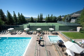 A pool area with a tennis court, lounge chairs, umbrellas, and a barbecue grill.
