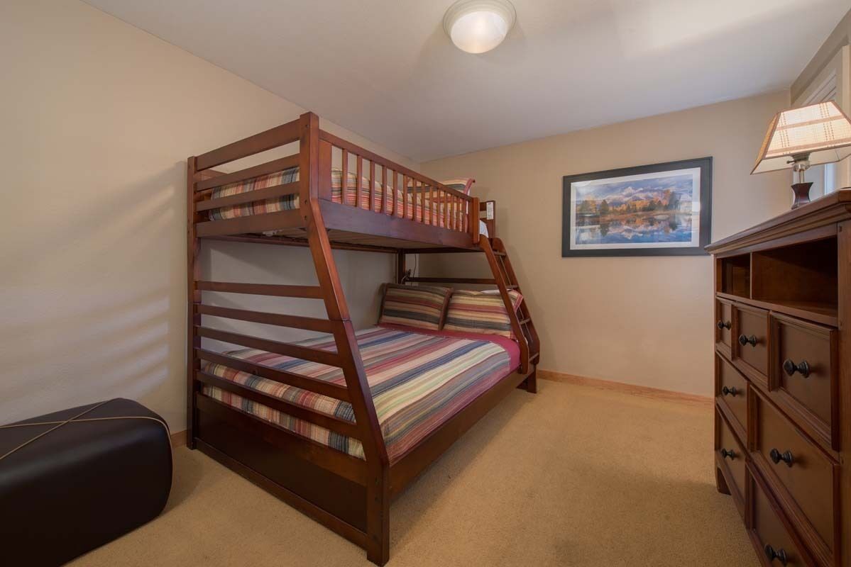 A bunk bed with a wooden frame and striped bedding, a black bench, a dresser with multiple drawers, and a framed picture on the wall.