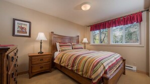 A bedroom with a wooden bed, two bedside tables, a nightstand, a window with red curtains, and a framed picture on the wall. - JHRL - Tensleep A2 - First floor condominium located in Teton Village (Teton Village)
