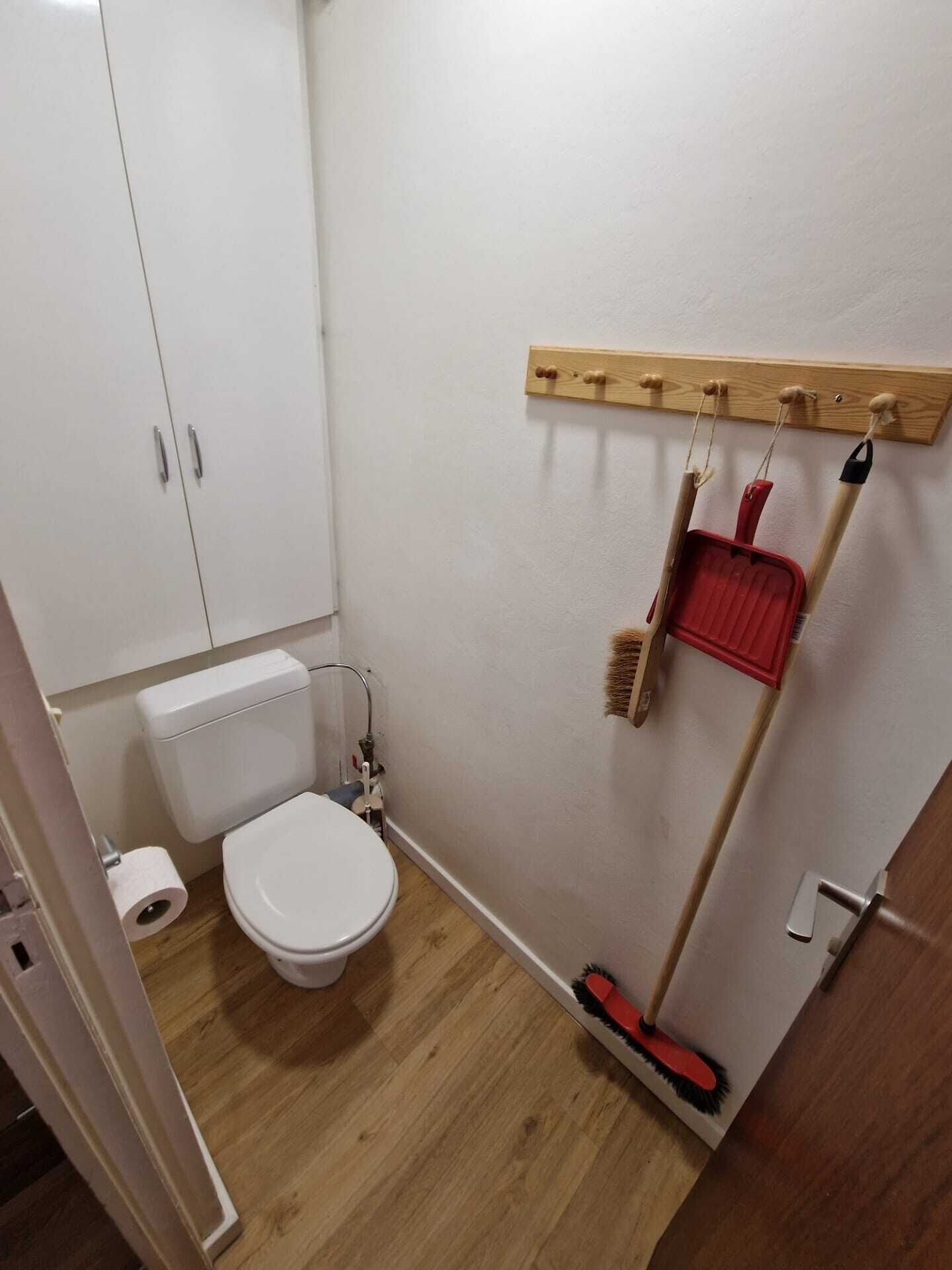 A small, clean bathroom with a toilet, a wooden floor, and a wall-mounted shelf holding cleaning tools.