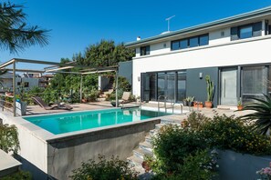 Une piscine extérieure moderne, dotée d’un bord en béton, entourée de plantes en pot et surplombée d’un ciel bleu azur.