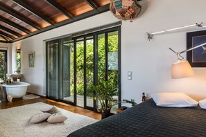 Une chambre avec un grand lit, une table de chevet, une plante en pot et une vue sur l’extérieur grâce à une porte coulissante.