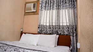 A bedroom with a bed, white pillows, a patterned bedspread, a window with floral curtains, and an air conditioning unit on the wall. - Maxton Suites Magodo (Lagos)