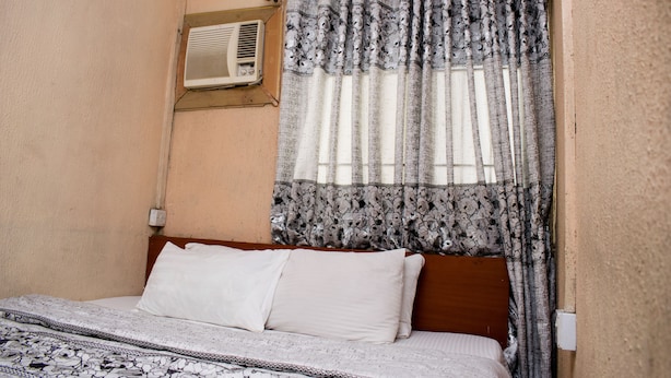 Un dormitorio con cama, almohadas blancas, colcha estampada, ventana con cortinas florales y un equipo de aire acondicionado en la pared.