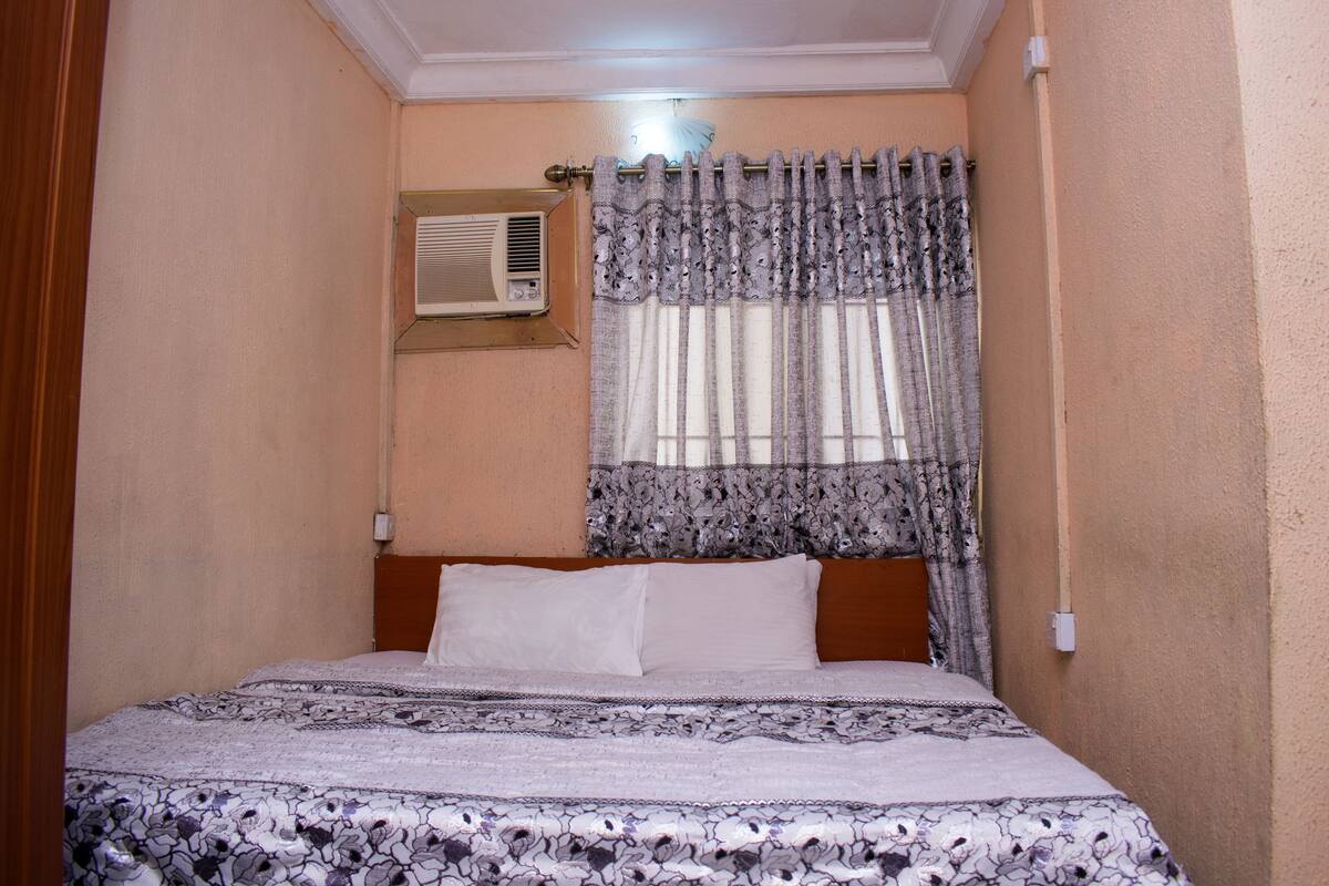 A bedroom with a bed, a window with patterned curtains, an air conditioning unit, and a wall-mounted light fixture.