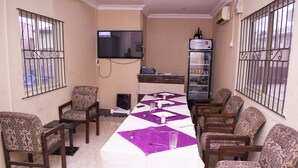 Un salón de conferencias con una larga mesa, sillas estampadas, un televisor y un refrigerador.