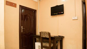 Una habitación con puerta de madera, televisor montado en la pared, un electrodoméstico pequeño y blanco, y una silla estampada.