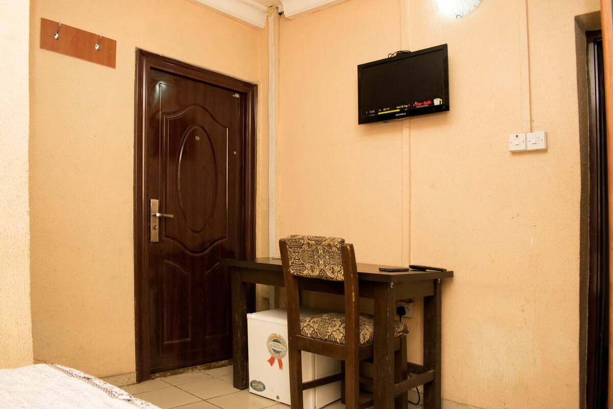 A room with a wooden door, a television mounted on the wall, a small white appliance, and a patterned chair.