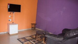A living room with a flat-screen TV mounted on the wall, a small white cabinet, a round glass table with two chairs, and a black leather sofa. - Emota Paradise Phase 2 (Lagos)