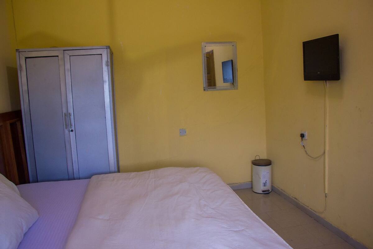 A room with a bed, a wardrobe, a mirror, a television, and a trash bin.
