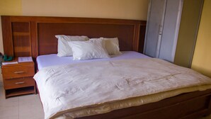 A neatly made bed with white linens and pillows, a wooden headboard, a wooden nightstand with drawers, and a wardrobe. - Emota Paradise Phase 2 (Lagos)