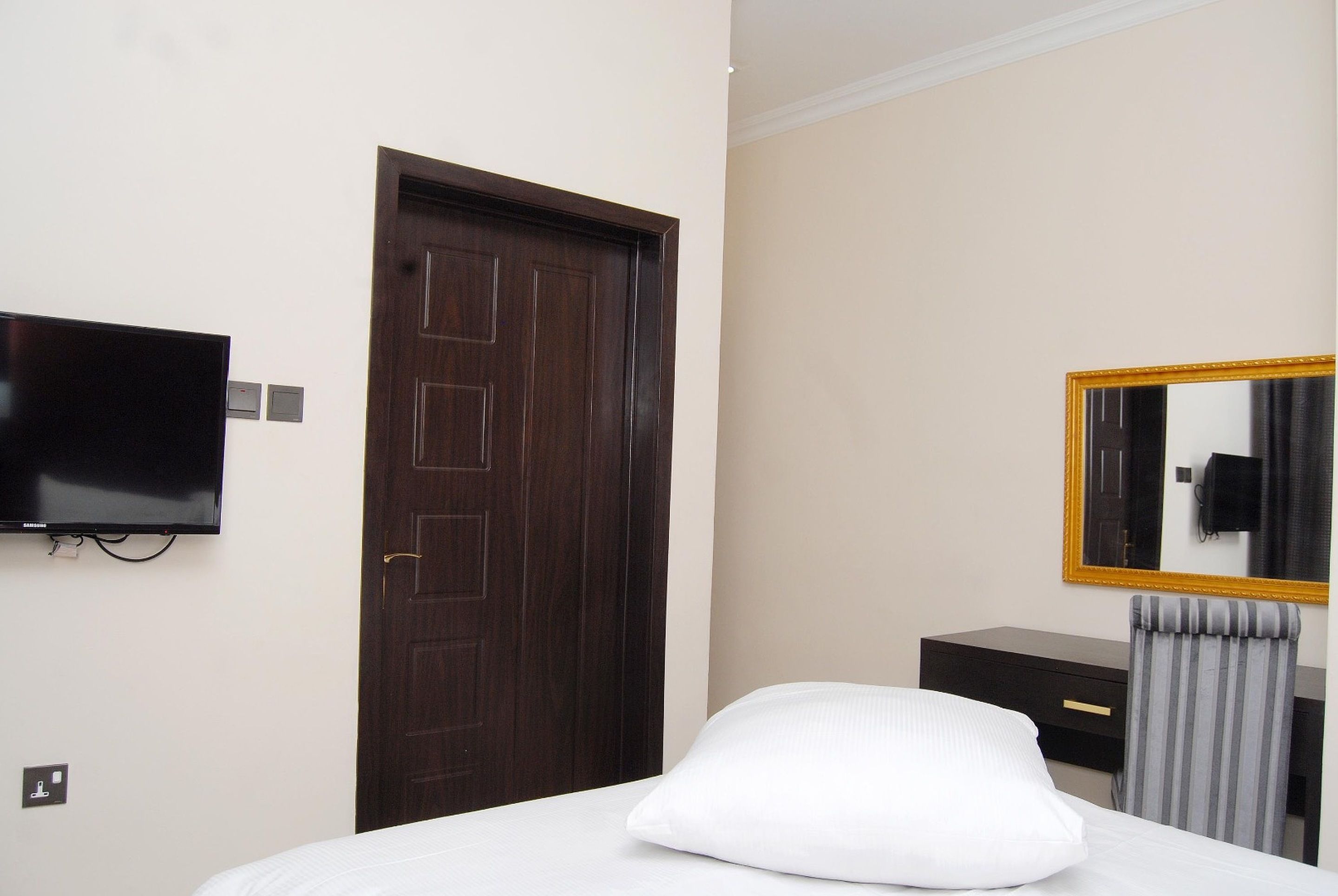 A hotel room with a bed, a television mounted on the wall, a wooden door, a dresser, and a mirror with a gold frame.