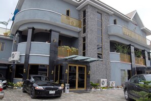 Un hotel de varios pisos con diseño moderno, que cuenta con balcones, una entrada central con puertas de vidrio y unidades de aire acondicionado.