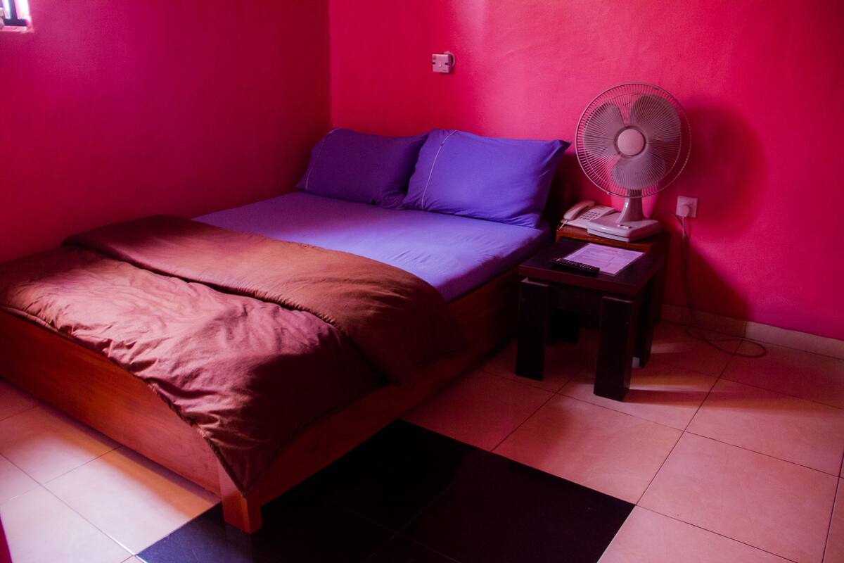 Ein Zimmer mit einem Bett, einem Nachttisch und einem Wandventilator.