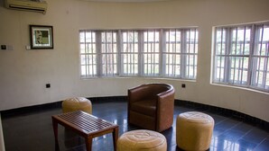 A room with a curved layout, featuring a brown leather chair, a wooden table, and two ottomans. Large windows provide natural light and a view of the outdoors. - Chelsea Suites Annex (Lagos)