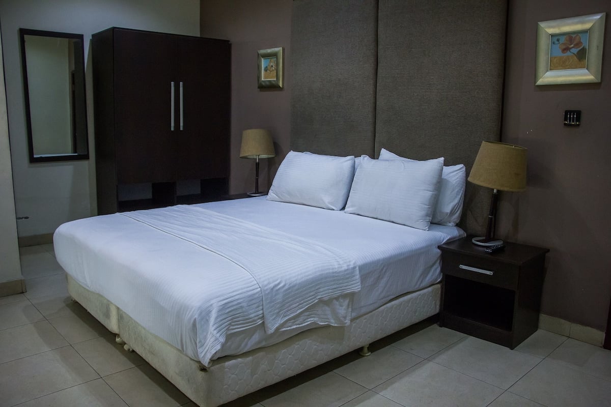 A neatly made bed with white linens, a dark wooden wardrobe, two bedside lamps, and a framed picture on the wall.