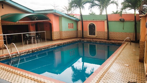 A swimming pool with a tiled deck, a covered seating area, and a building with a green roof and brick walls. - Alfoscar Hotel & Suites (Lagos)