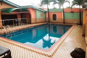 A swimming pool with a tiled deck, a covered seating area, and a building with a green roof and brick walls. - Alfoscar Hotel & Suites (Lagos)