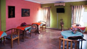 A dining area with wooden chairs and tables, a flat-screen TV mounted on the wall, and a window with striped curtains. - Alfoscar Hotel & Suites (Lagos)
