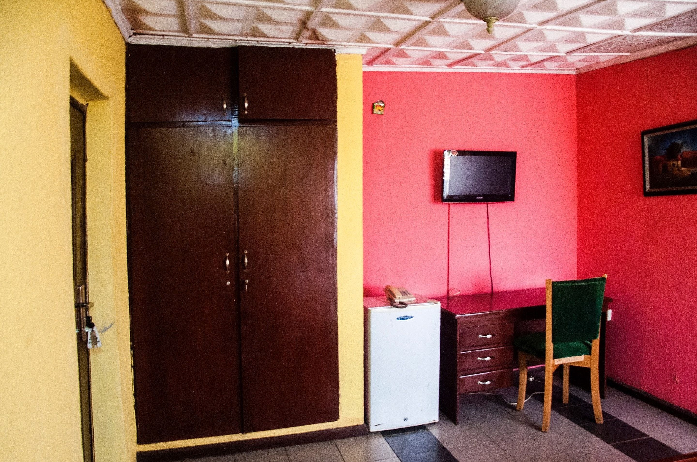 A room with a brown wardrobe, a small white refrigerator, a desk with a chair, a television mounted on the wall, and a red wall.